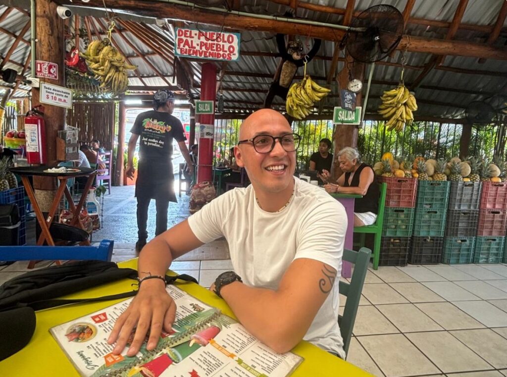 Habibi Yassine sitting at a table in Restaurante Nativo, smiling and looking to the side, with a menu in front of him and hanging bunches of bananas and tropical decor in the background.