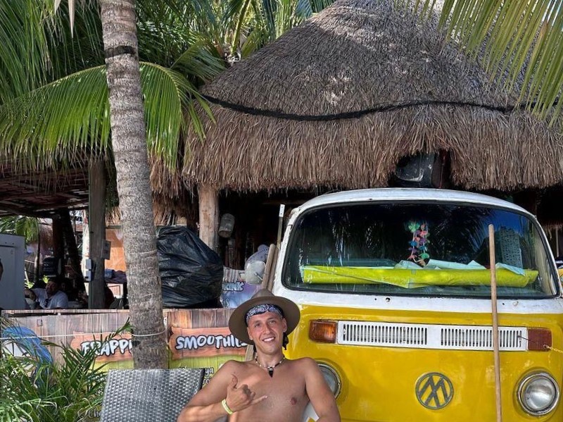 Habibi Yassine wearing a hat and a bandana, sitting in front of a yellow Volkswagen van with a thatched roof hut and palm trees in the background on Isla Mujeres.