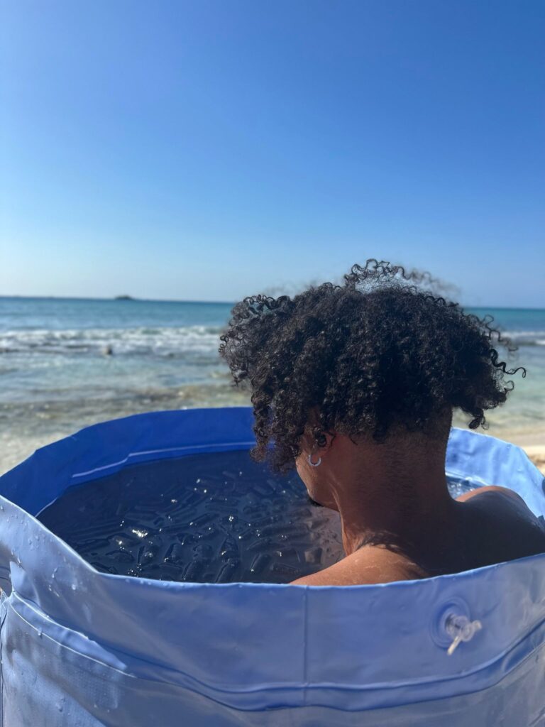 Person with curly hair sitting in an ice bath on the beach, facing the ocean under a clear blue sky.